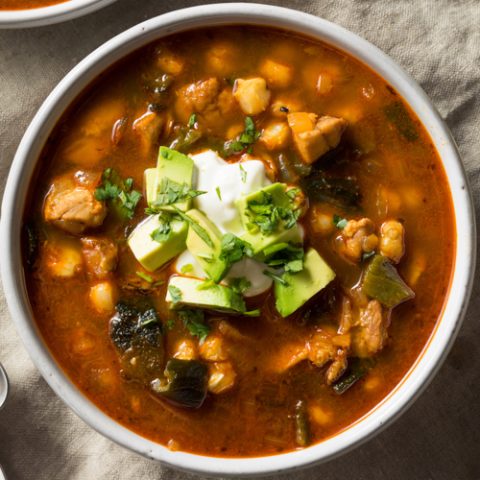 bowl of Mexican soup called pozole in white bowls garnished with sour cream and avocado