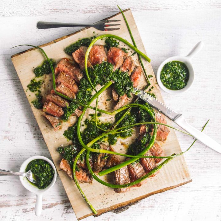 Grilled steak with salsa verde on a cutting board with small bowls of more salsa verde on the side, on a white background.