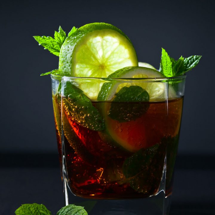 Cuba libre cocktail in a glass garnished with lime on a black background.