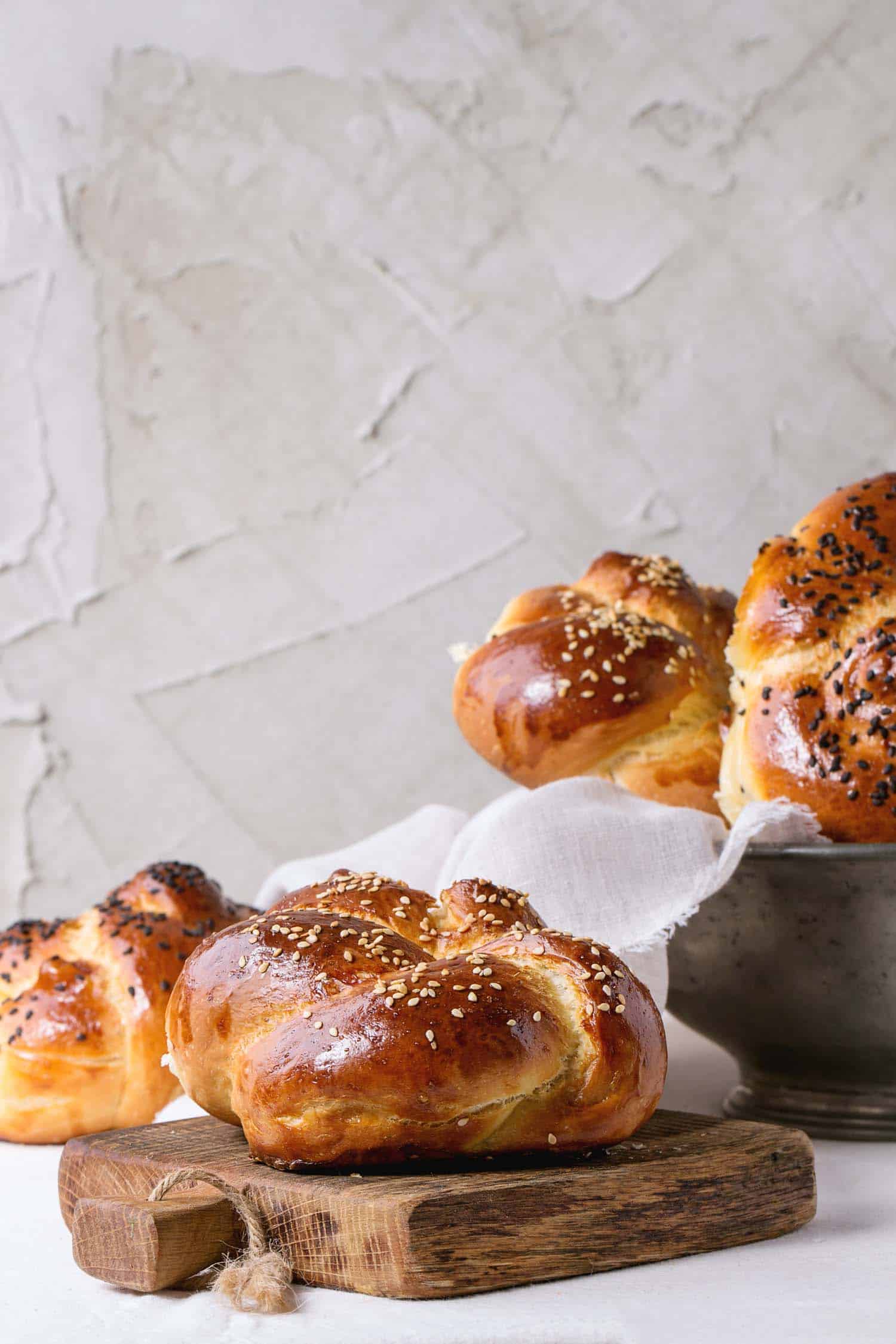 Heap of sweet round sabbath challah bread, traditional Rosh Hashanah food with white and black sesame seeds in vintage metal bowl and on small cutting board over white table with plastered wall at background.