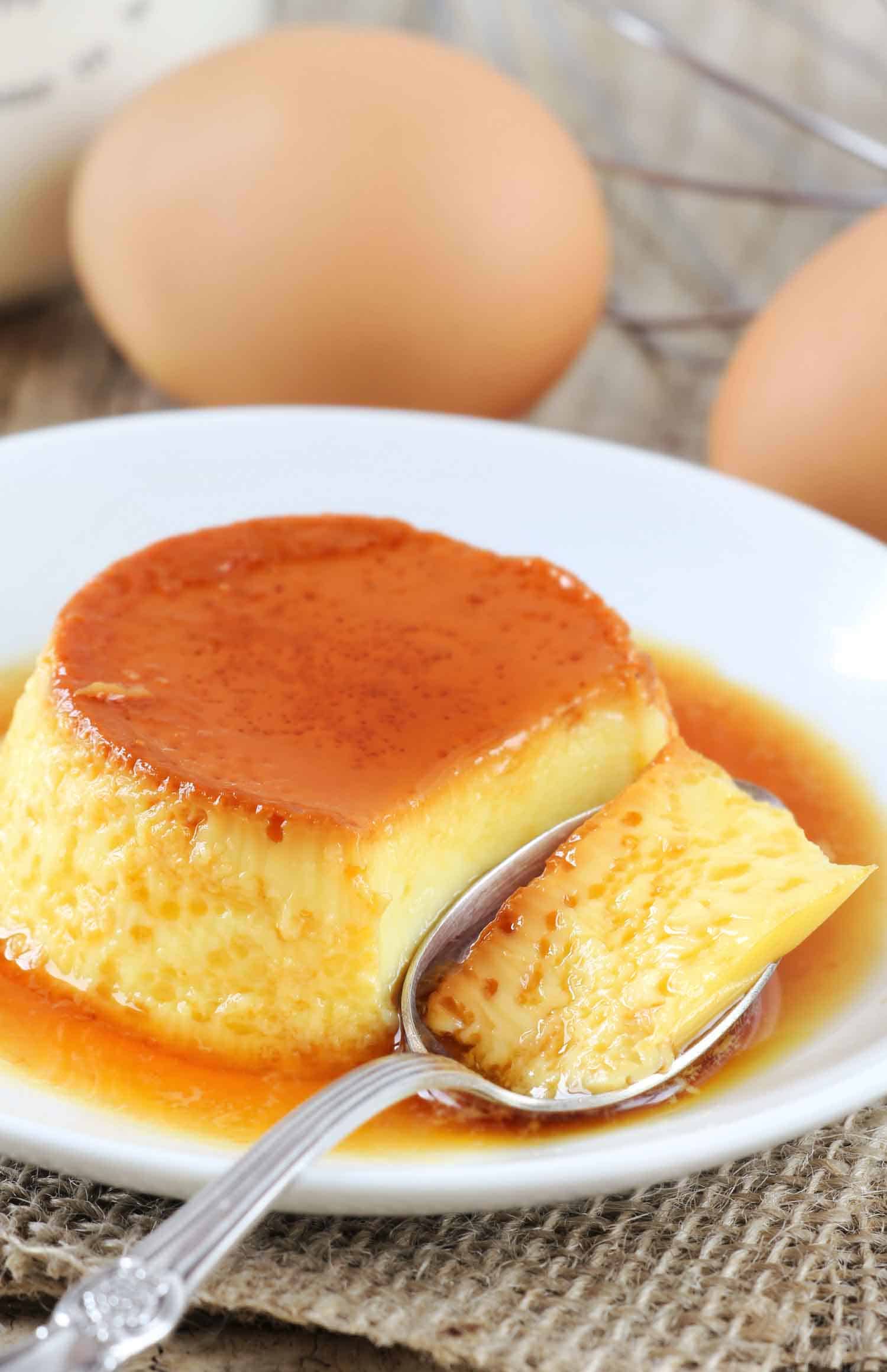 Flan that has been cut into with a spoon on a white plate with eggs in the background.