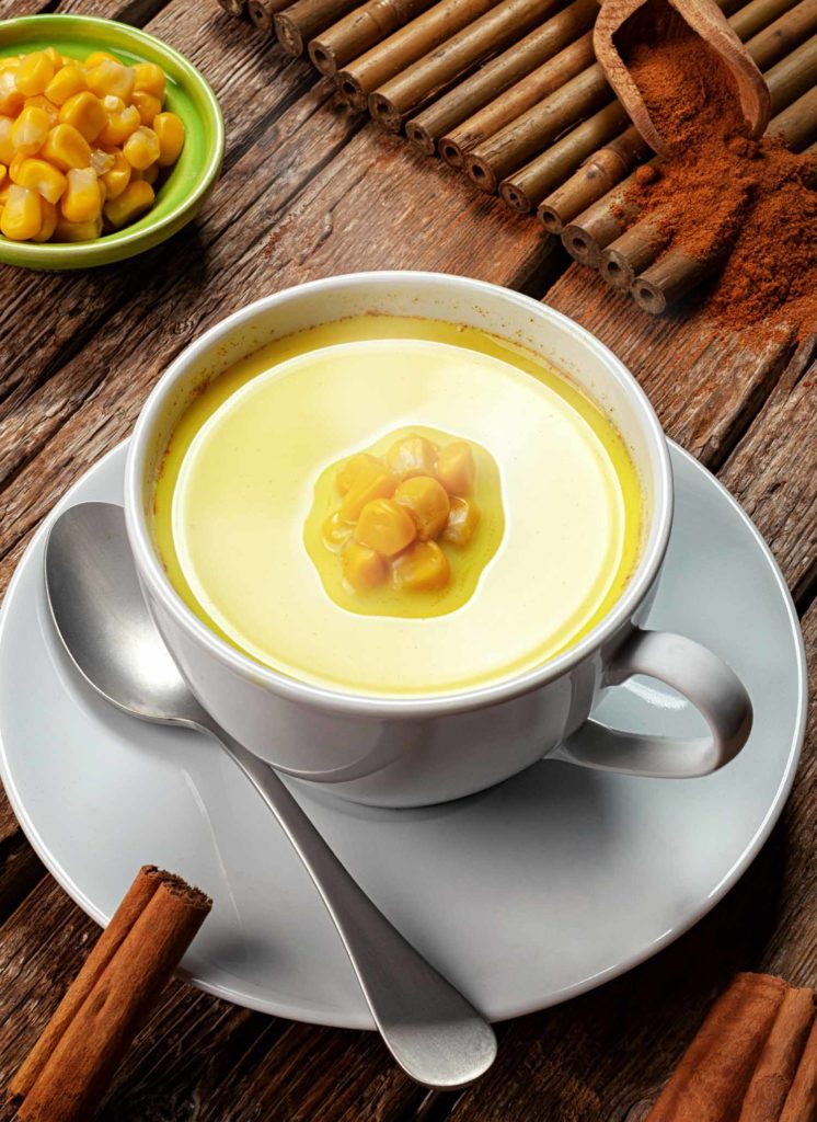 Atol de elote, one of the most common Honduran drinks, on a table with corn and cinnamon garnishes.