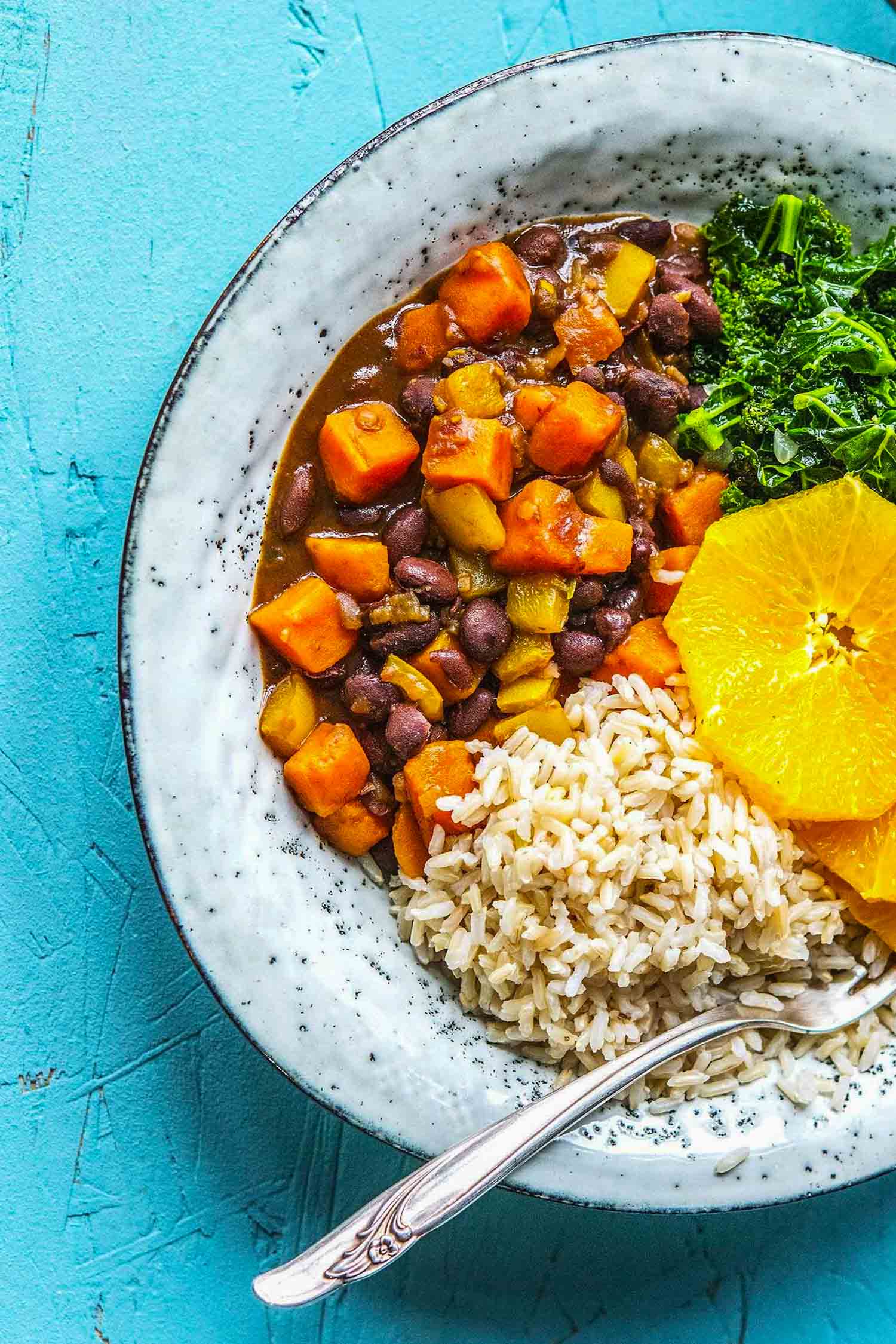 Vegetarian feijoada, a black bean stew with squash on white plate with kale and sliced oranges on a turquoise background.