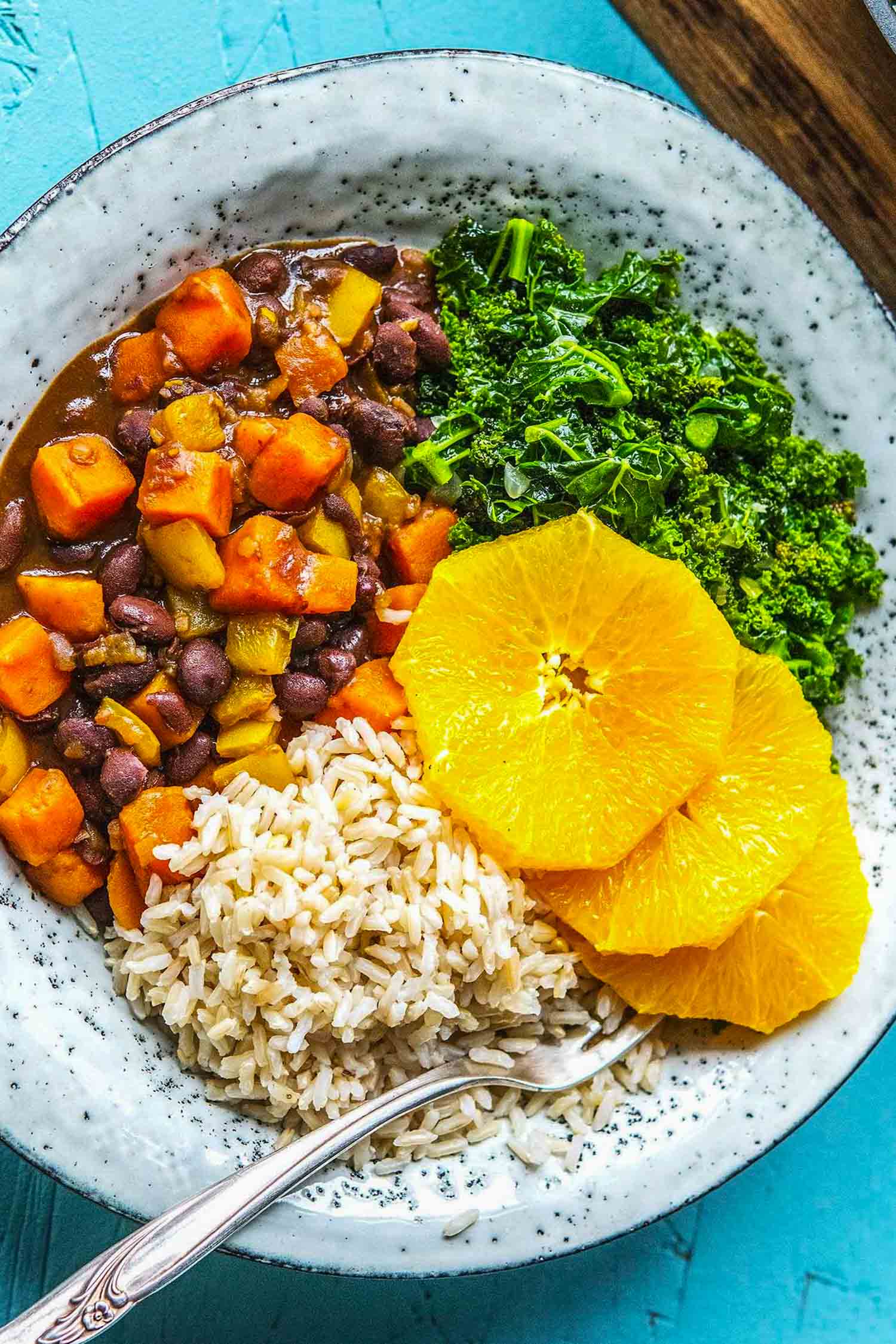 Vegetarian feijoada, a black bean stew with squash on white plate with kale and sliced oranges on a turquoise background.