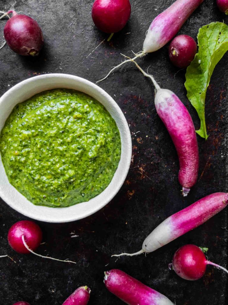 Radish top pesto on a dark background surrounded by two kinds of radishes and leaves