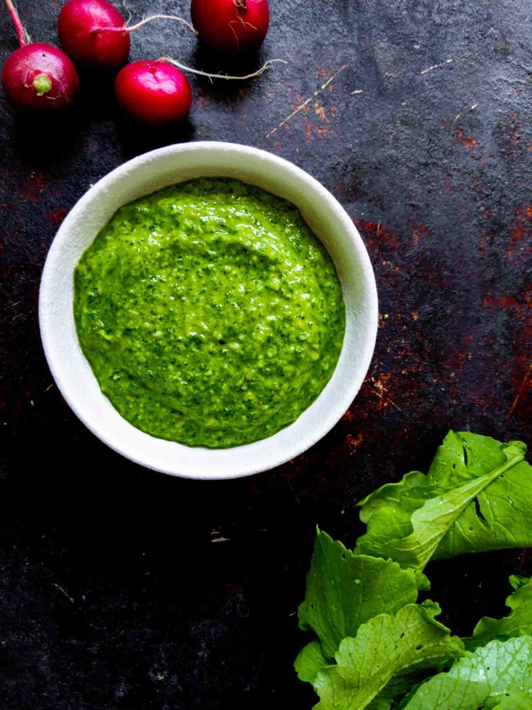 White bowl with radish greens pesto on dark background with radishes and radish leaves in corners