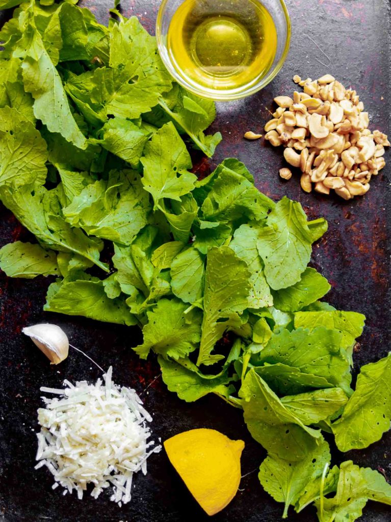 Ingredients to make radish greens pesto on dark background, small bowl of canola oil, cashews, radish leaves, close of garlic, shredded parmesan cheese and a quarter of a lemon