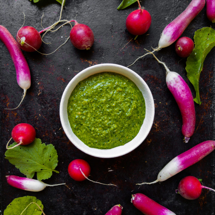 Radish green pesto on a dark background surrounded by two kinds of radishes and leaves