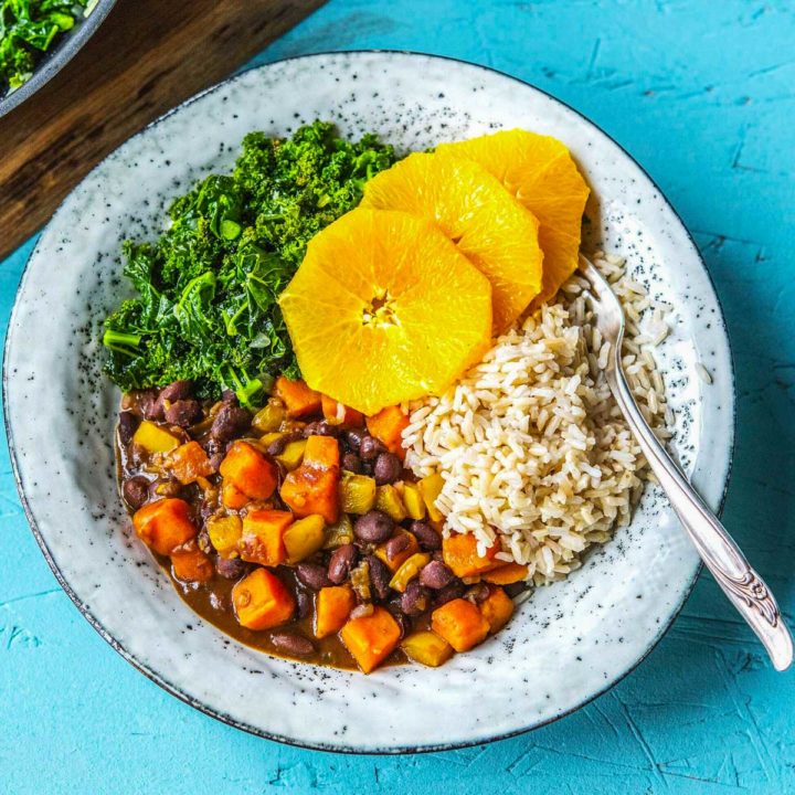 Vegetarian feijoada, a black bean stew with squash on white plate with kale and sliced oranges on a turquoise background.