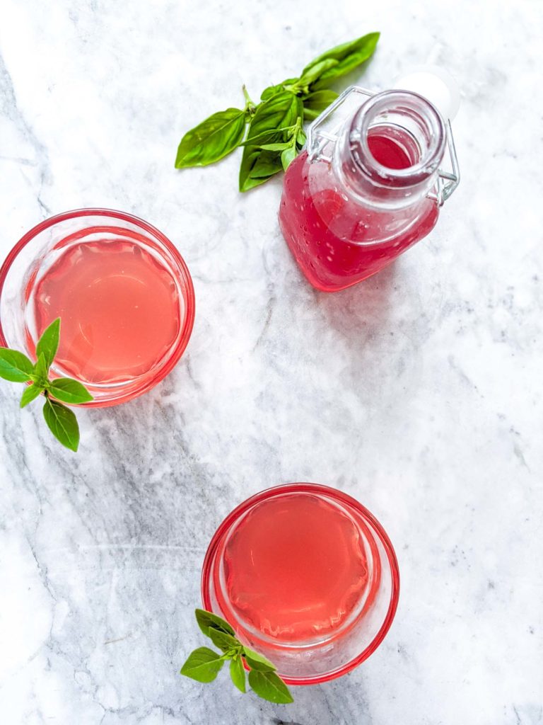Flatlay view of two rhubarb cocktails with bottle of rhubarb syrup garnished with basil