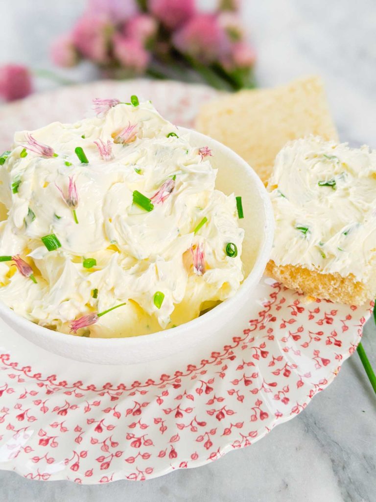 chive butter in a white bowl on a plate alongside two pieces of buttered bread with chives behind it on a white background