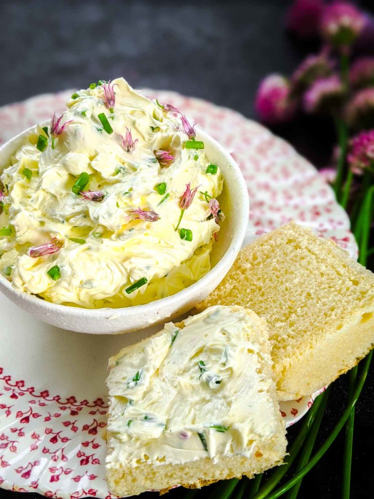 chive butter in a mason jar on a dark background in a bowl with two pieces of bread on a plate and chives with blossoms in the background