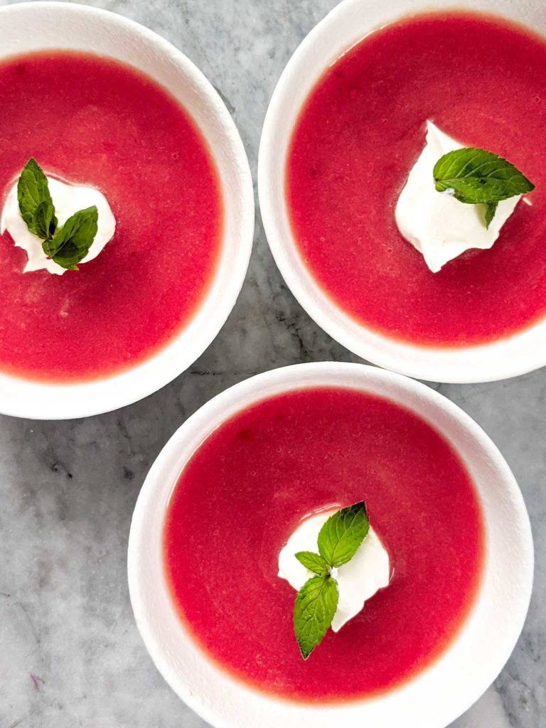 Cold rhubarb soup topped with whipped cream and mint in three white bowls on a grey marble background.