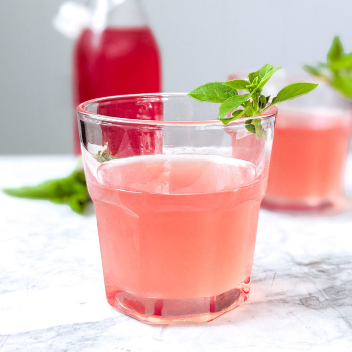 Basil rhubarb shrub in bottle next to two cocktails using it, garnished with basil.