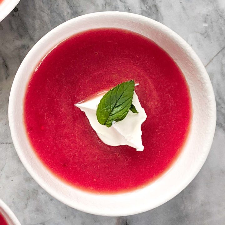 Cold rhubarb soup in a white bowl, garnished with mint and cream on a white marble background.