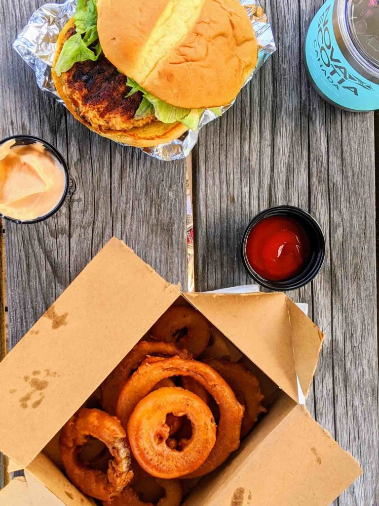 Top view of food on table at Inverness Beach Hut on wooden picnic table with crab cake sandwich, onion rings, ketchup and sriracha mayo.