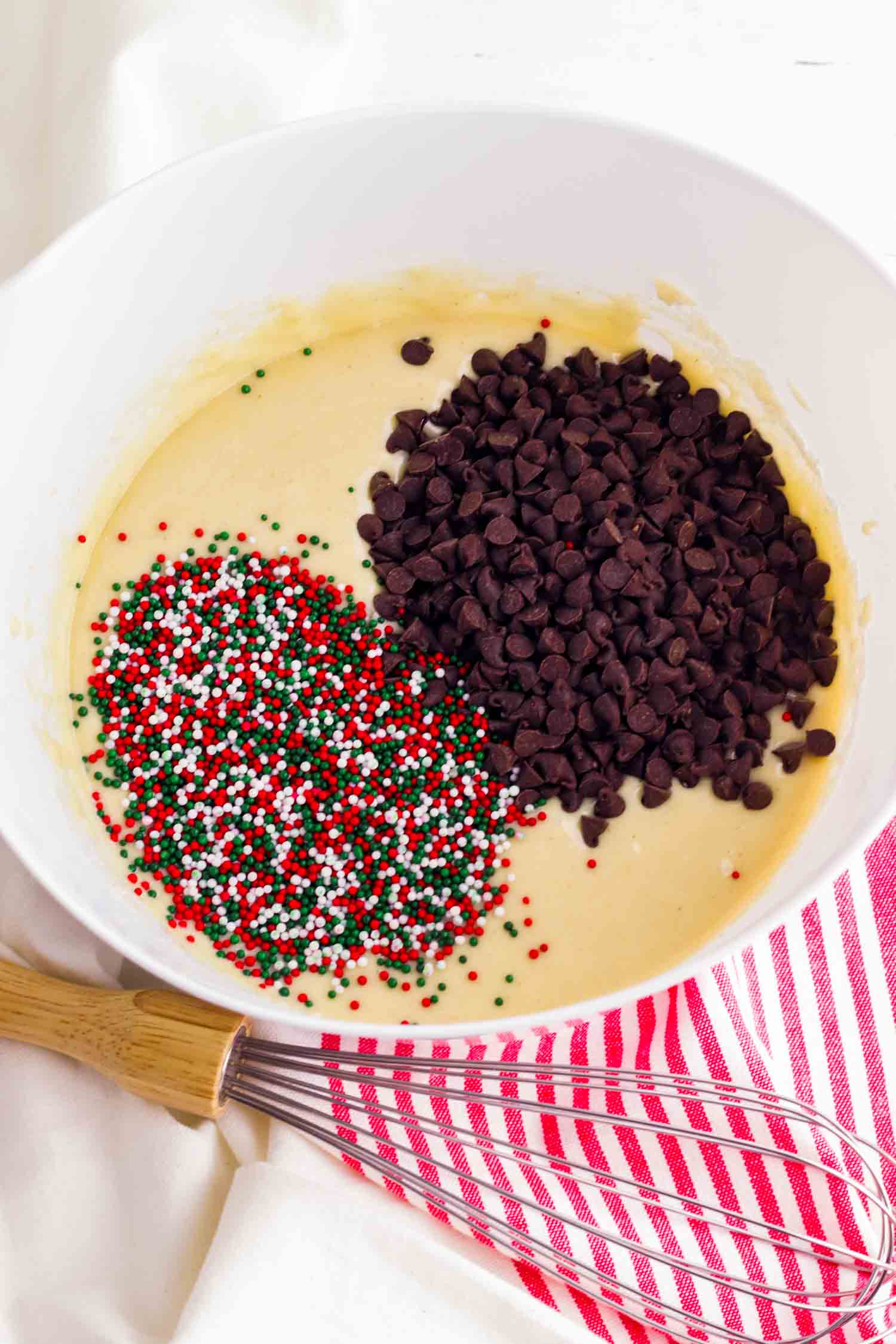 Mini chocolate chips and Christmas sprinkles in white bowl with whisk and red and white tea towel.