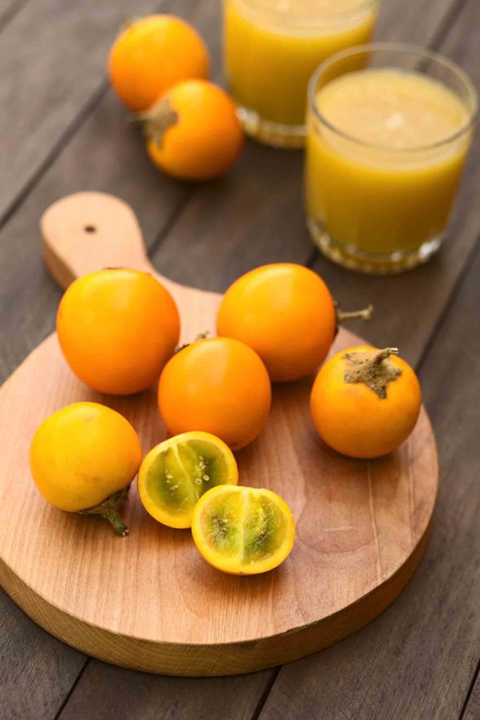 Naranjilla or Lulo fruits (lat. Solanum quitoense) on wooden board with freshly prepared naranjilla juice in the back (Selective Focus Focus on the first naranjilla half)