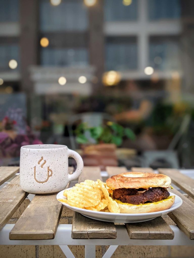 Fredericton Restaurants The Tipsy Muse Impastrami vegan pastrami on rosemary sea salt bagel on table with coffee on patio