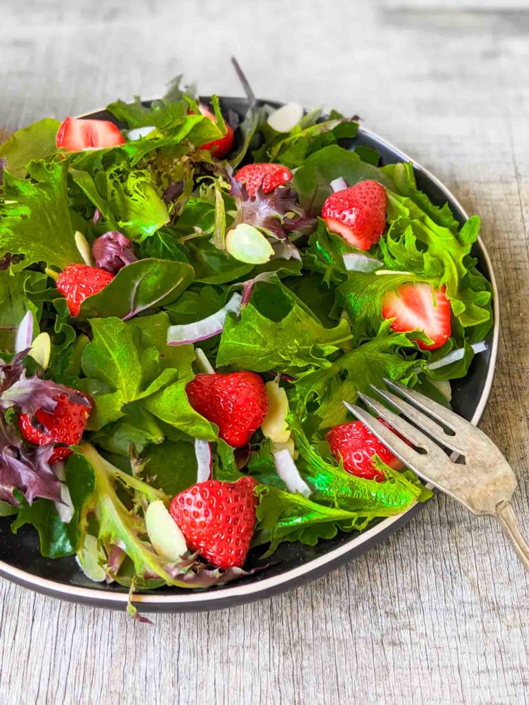 Strawberry salad with spinach, spring mix, arugula, red onion, slivered almonds on a black plate and rustic surface.