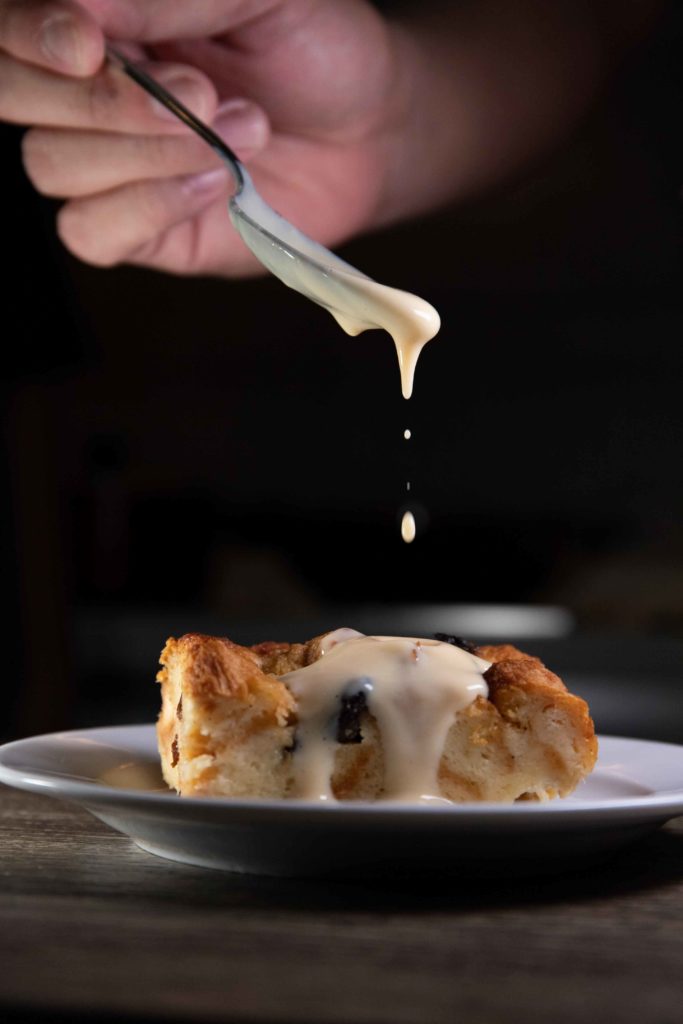 Budin is a common dessert in Costa Rica known as bread pudding, it is on a white plate with a hand adding a sweet topping