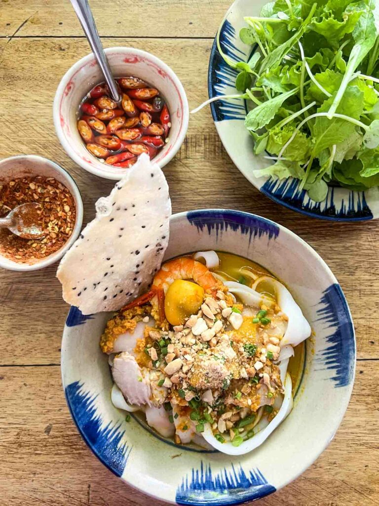 mi quang noodles in Hoi An Vietnam restaurant, features traditional recipe with pork, shrimp, quail egg alongside bowl of greens, chili vinegar and chili flakes